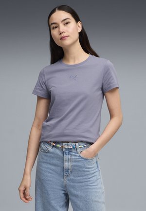 Mujer de cabello largo y oscuro que lleva una camiseta Puma morada clara y jeans azul claro con un cinturón colorido de cuentas, sobre un fondo gris.