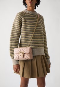 Mīksts rozā kviltots crossbody soma ar zelta apaļu aizdari, ar ķēdes siksnu. Modelle valkā svītrainu džemperi un olīvzaļas krāsas svārkus.