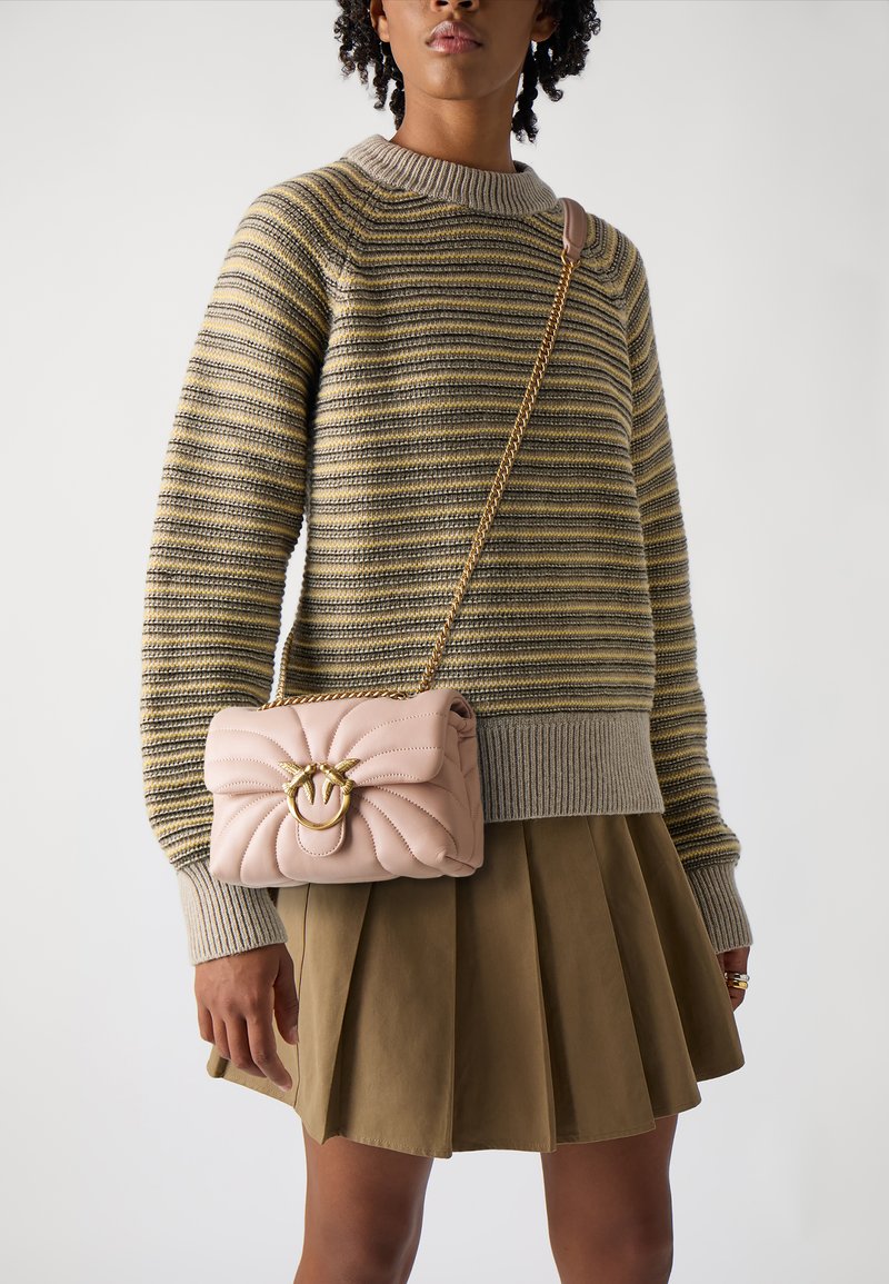 Mīksts rozā kviltots crossbody soma ar zelta apaļu aizdari, ar ķēdes siksnu. Modelle valkā svītrainu džemperi un olīvzaļas krāsas svārkus.