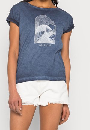 Femme portant un t-shirt bleu Roxy avec un motif blanc de vague et de soleil, associé à un short en jean blanc effiloché.