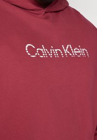 Bordowa bluza z kapturem wykonana z miękkiej mieszanki bawełny; posiada biały napis "Calvin Klein" w kropkowanym wzorze. Prosty, luźny fason.
