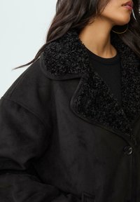 Crna lažna antilop jakna s teksturiranim crnim krznom na ovratniku, s jednim gumbom za zatvaranje i širokim, opuštenim krojem.