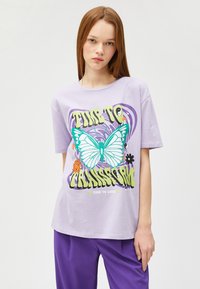 Camiseta de manga corta en color lila con un gran estampado gráfico de una mariposa y el texto "ES HORA DE TRANSFORMARSE", con acentos coloridos.
