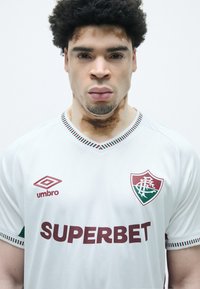 Camisola de futebol branca com um emblema vermelho e verde, logo "SUPERBET" em bordô, e detalhes em listras nas mangas e colarinho.