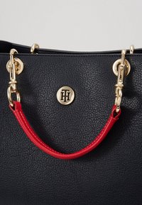 Granatowa skórzana torebka z gładką fakturą, złotymi elementami i kontrastującym czerwonym uchwytem z okrągłym logo z przodu.