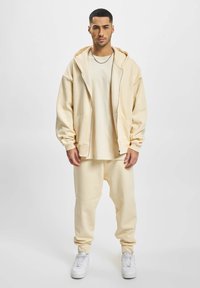 Beige zip-up hoodie med store lommer, parret med matchende joggers og hvide sneakers. Enkel design, blød tekstur og afslappet pasform.