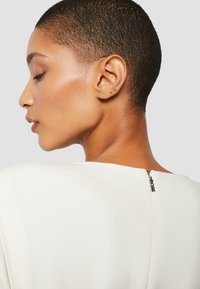 Witte top met een ronde halslijn, voorzien van een ritsdetail aan de achterkant. Korte, nauw sluitende haarsnit en gouden stud oorbellen zijn zichtbaar.