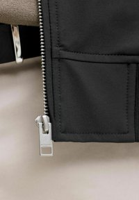 Gros plan d'une fermeture éclair argentée partiellement ouverte sur un tissu noir avec une poche carrée cousue et une surface beige texturée en arrière-plan.