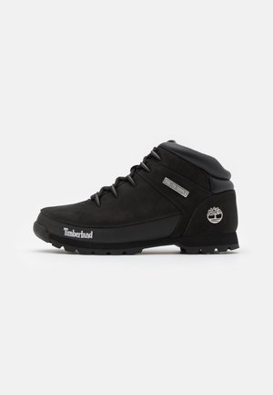 Schwarze wasserdichte Wanderschuhe mit einem Obermaterial aus Wildleder und Synthetik, Gummisohle, gepolstertem Schaft und metallic Logo an der Ferse. Mit Schnürung.