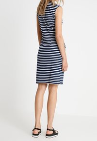 Kvinna som står, klädd i en ärmlös marinblå och vit randig knälång klänning med svarta sandaler mot en enfärgad bakgrund.