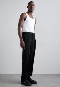 Černé kalhoty se střihaným střihem, doplněné bílým tílkem. Model nosí černé tenisky s texturovanými tkaničkami.