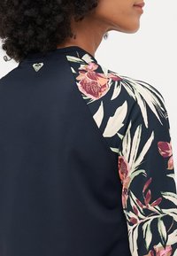 Haut de sport marine avec des manches à motif floral dans les tons de rose, vert et blanc. Logo distinctif au dos du cou. Tissu lisse et extensible.