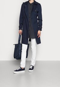 Navy trench coat with button closure, worn over a patterned shirt. White pants and navy sneakers complement the look. Includes a matching tote bag.