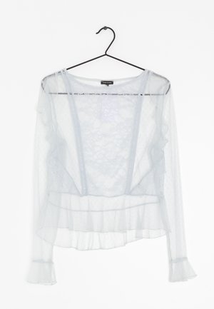 Blouse blanche transparente à manches longues avec des détails en volants sur les manches et l'ourlet, motif floral, bande de dentelle verticale, suspendue sur un cintre noir.