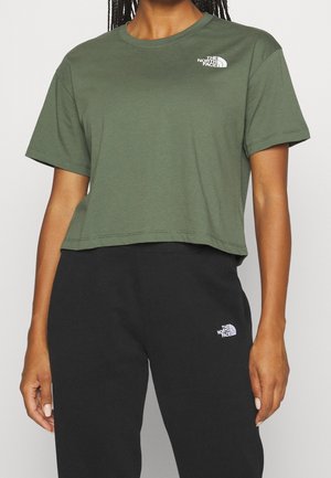 Kobieta w oliwkowym, krótkim T-shircie i czarnych dresach, oba z logo The North Face na piersi i udzie.