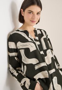 Černá a béžová halenka s abstraktním vzorem štětcových tahů. Vyrobena z lehké látky, má výstřih bez límce a dlouhé rukávy.