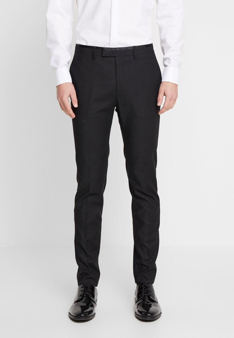 Svarta skräddarsydda byxor med en mjuk tygfinish, smal passform och subtila veck framtill, tillsammans med glänsande svarta kostymskor.