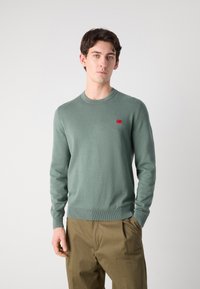Light green knitted sweater with a round neckline and long sleeves, featuring a small red logo on the chest and ribbed cuffs and hem.