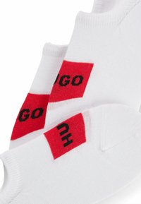 Weiße Knöchel-Socken mit roten und schwarzen Logoakzenten. Das Logo erscheint in fetter Schrift auf einem roten rechteckigen Patch. Weiches, atmungsaktives Material.