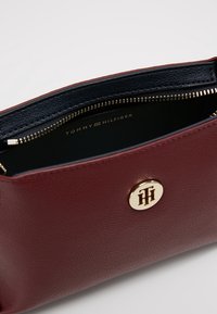 Bolso de cuero burdeos con herrajes en tono dorado, cierre con cremallera y un interior azul marino. Presenta el logo de Tommy Hilfiger grabado en el interior.
