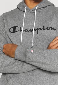 Felpa grigia realizzata in morbido tessuto con il logo "Champion" nero. Presenta cordini bianchi e un piccolo emblema rosso, bianco e blu sul polsino.