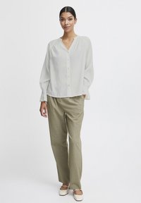Blusa grigia chiaro con scollatura a V e chiusura a bottoni, abbinata a pantaloni beige. La blusa presenta maniche a sbuffo con polsini elastici.