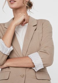 Jasnobeżowa marynarka o dopasowanym kroju, z zapięciem na jeden guzik i podwiniętymi białymi rękawami w paski dla kontrastu.
