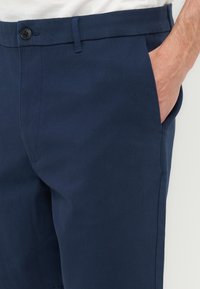 Άνδρας που φοράει σκούρο μπλε παντελόνι με το χέρι μέσα στην δεξιά μπροστινή τσέπη, ορατή η μέση με κουμπιά.