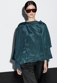 Sjøgrøn bluse i silke med et asymmetrisk design, lagdelte folder og brede ærmer, parret med sorte bukser. Modellen bærer mørke, overdimensionerede solbriller.