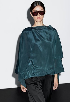 Türkise Seidenbluse mit asymmetrischem Design, geschichteten Falten und weiten Ärmeln, kombiniert mit schwarzen Hosen. Das Model trägt dunkle, übergroße Sonnenbrillen.