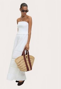 Witte strapless maxi-jurk van gestructureerde stof met kanten accenten, gecombineerd met een strooien tas met bruine band en donkere platte schoenen.