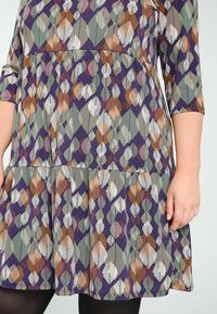 Robe violette avec une jupe à volants, présentant un motif géométrique de feuilles en vert, blanc, orange et marron. Manches trois-quarts et texture douce.