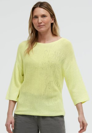 Woman wearing a light yellow knit sweater with 3/4 sleeves and loose fit, paired with gray pants, standing against a plain light background.
