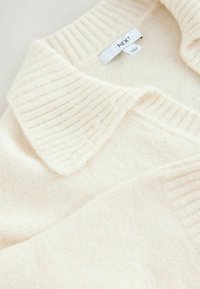 Pull crème avec un col côtelé et un tissu texturé. Présente une étiquette avec "NEXT." Texture lisse et apparence douce et chaleureuse.