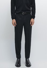 Mand iført tætsiddende sorte bukser, sort læderbælte, sort langærmet skjorte og sorte læderstøvler mod en ensfarvet baggrund.