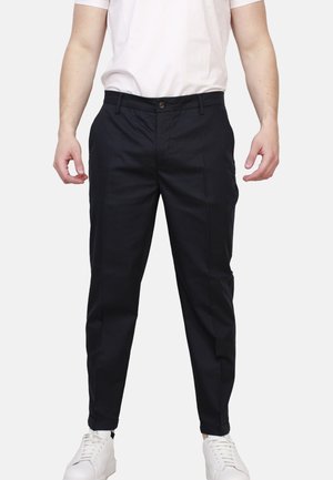 Uomo con pantaloni neri aderenti, sneaker bianche e t-shirt bianca infilata, in piedi con le braccia rilassate ai lati.