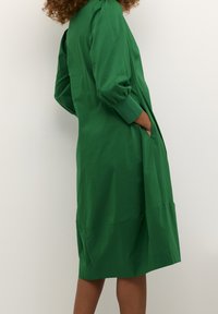 Grön, knälång klänning i slätt tyg med långa ärmar, puffiga axlar, sidfickor och en enkel passform.