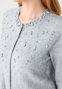 Grijze cardigan van zachte stof met versieringen van heldere strass-steentjes en kristallen in verschillende vormen over de voorkant en halslijn.