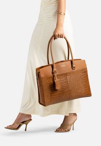 Sac fourre-tout en cuir marron avec texture crocodile, poignées doubles et fermeture éclair. Étiquette avec logo incluse. Design lisse et structuré.