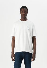 White cotton t-shirt with short sleeves, a crew neck, featuring a small blue graphic and logo on the chest. Denim jeans worn below.
