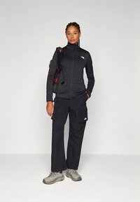 Ženska stoji oblečena v črno jakno z zadrgo, črne delovne hlače, sive pohodniške čevlje in ima nahrbtnik čez eno ramo.