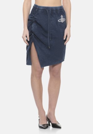 Donna che indossa una gonna blu navy al ginocchio con spacco arricciato laterale, vita con coulisse, patch con logo e tacchi neri a punta.