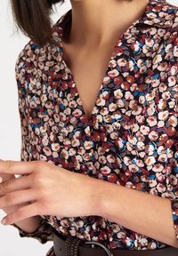 Blouse florale avec un fond sombre ornée de petites fleurs roses, blanches et bleues ; design à col ; tissu léger ; détail à boutons au niveau du cou.
