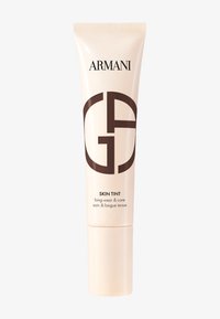 Beige tube av Armani Skin Tint med mørk brun logo, sylinderform og blank finish. Teksten inkluderer "langvarig og pleie."