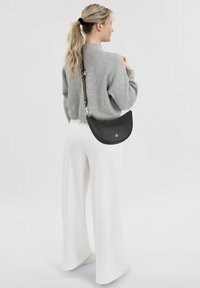 Bolso de cuero negro con forma curva, que cuenta con una correa desmontable con acentos metálicos. Llevado sobre un suéter gris y pantalones anchos blancos.