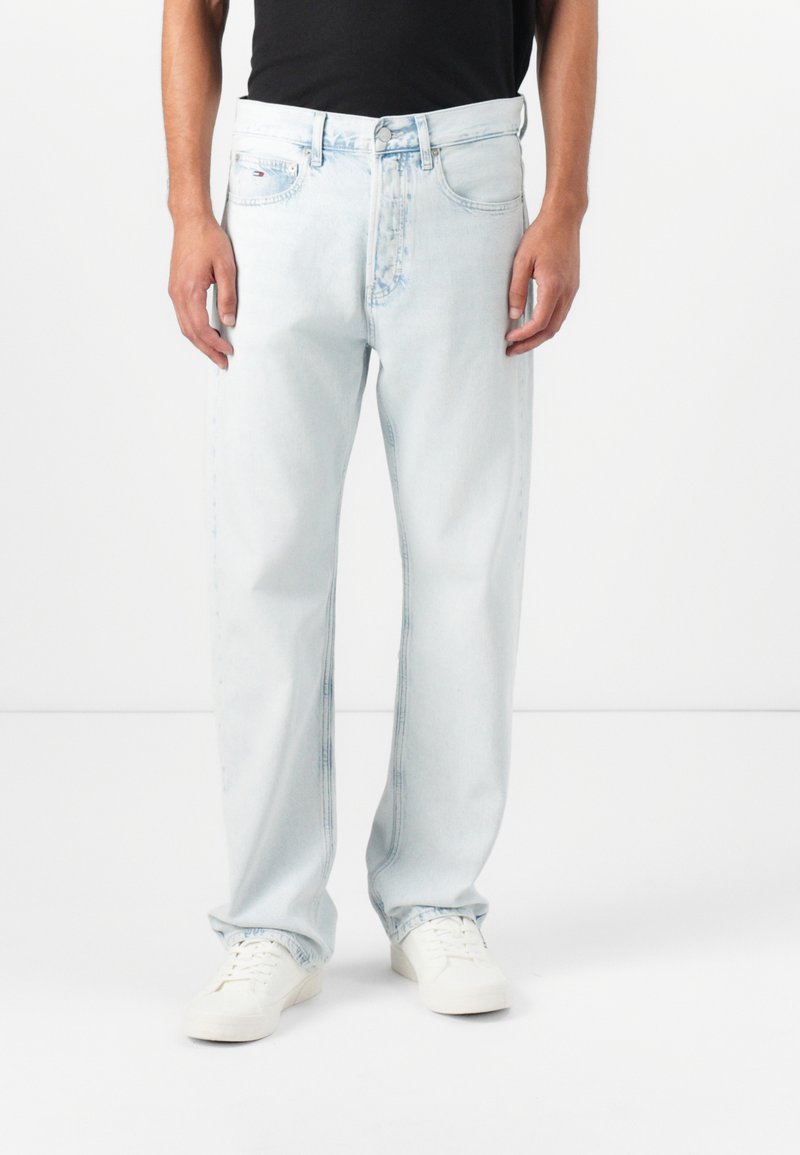 Jean en denim bleu clair avec une coupe droite, fermeture à bouton et poches avant, associés à des baskets blanches. Texture lisse.