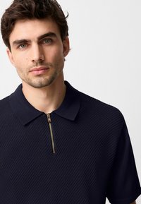 Polo shirt blu scuro con colletto con zip, caratterizzato da un motivo testurizzato e maniche corte. La zip dorata aggiunge un accento a contrasto.