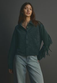 Dunkelgrünes Wildlederhemd mit Fransenverzierungen an den Ärmeln, großem Kragen und Knopfverschluss. Kombiniert mit hellblauen Jeans.