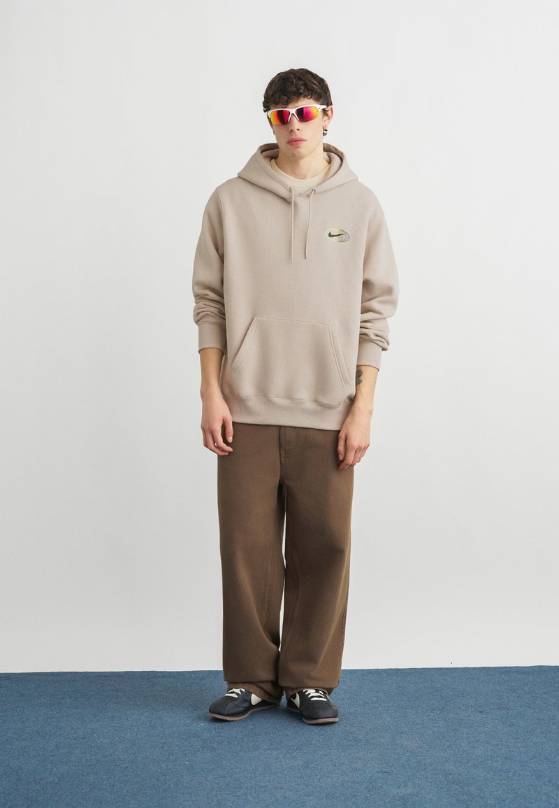 Giovane uomo con felpa con cappuccio Nike beige, pantaloni marroni ampi, scarpe da ginnastica nere e occhiali sportivi riflettenti rossi, in piedi su un tappeto blu.