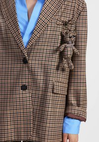 Blazer a quadri marrone con bottoni neri, dotato di una tasca e una camicia blu sotto. Un piccolo portachiavi a forma di orsacchiotto è attaccato al risvolto.
